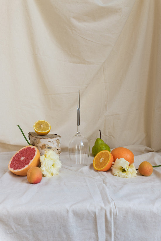 picnic wine glass with metal pin placed on a beige decor cloth next to halved orange, lemon and grapefruit, pear, apricots and white flowers