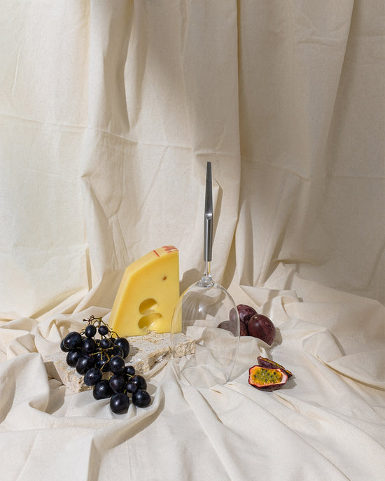 picnic wine glass for red wine with metal pin, placed upside down on a beige cloth decor next to a piece of Emmentaler cheese, a red grape and 2 plums
