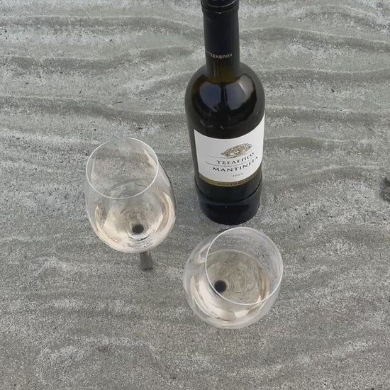 two beach wine glasses with a pointy metal pin placed in the sand and in the sea water next to a white wine bottle in Greek Island Paros