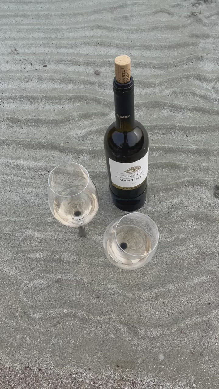 two beach wine glasses with a pointy metal pin placed in the sand and in the sea water next to a white wine bottle in Greek Island Paros