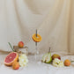 picnic wine glass with metal pin, placed upside down on a table cloth next to a halved grapefruit, halved pear, halved orange pierced by the metal pin, some apricots and flowers