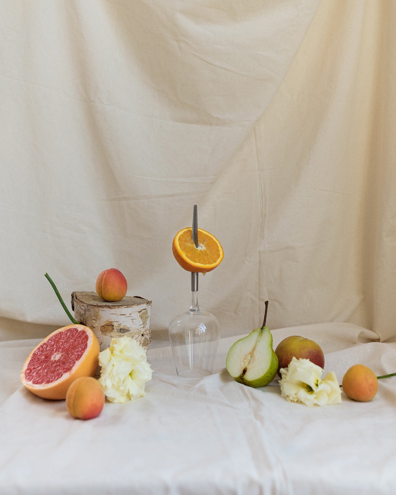 picnic wine glass with metal pin, placed upside down on a table cloth next to a halved grapefruit, halved pear, halved orange pierced by the metal pin, some apricots and flowers